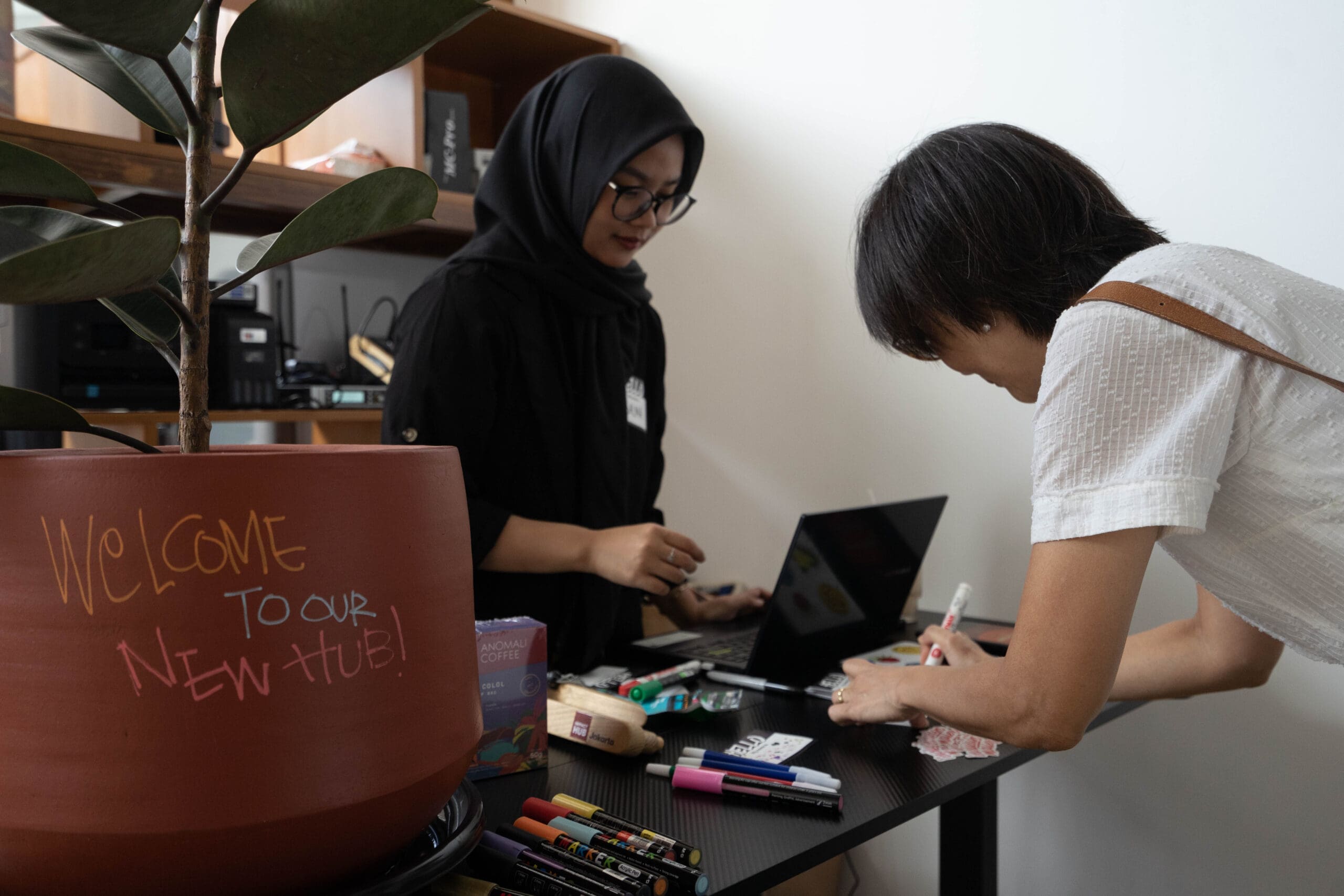 Two people at a membership registration table inside Impact Hub Jakarta, one assisting behind a laptop while the other fills out a form, with a “Welcome” sign and supplies visible on the table.