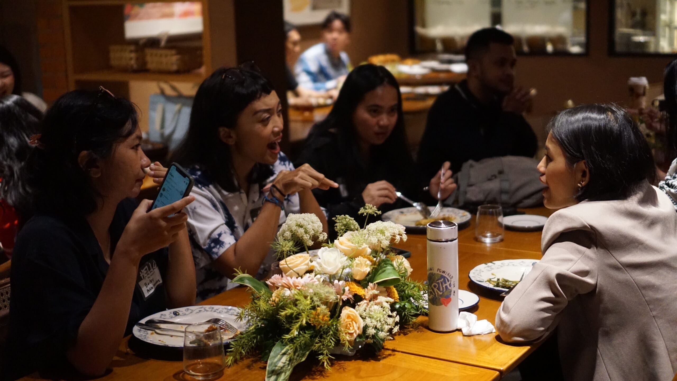 A group of people sitting around a long dining table, chatting and eating together, with floral table decorations and plates of food, capturing a lively networking and community moment during Bayer Women Empowerment Program Community Gathering that Impact Hub Jakarta held.