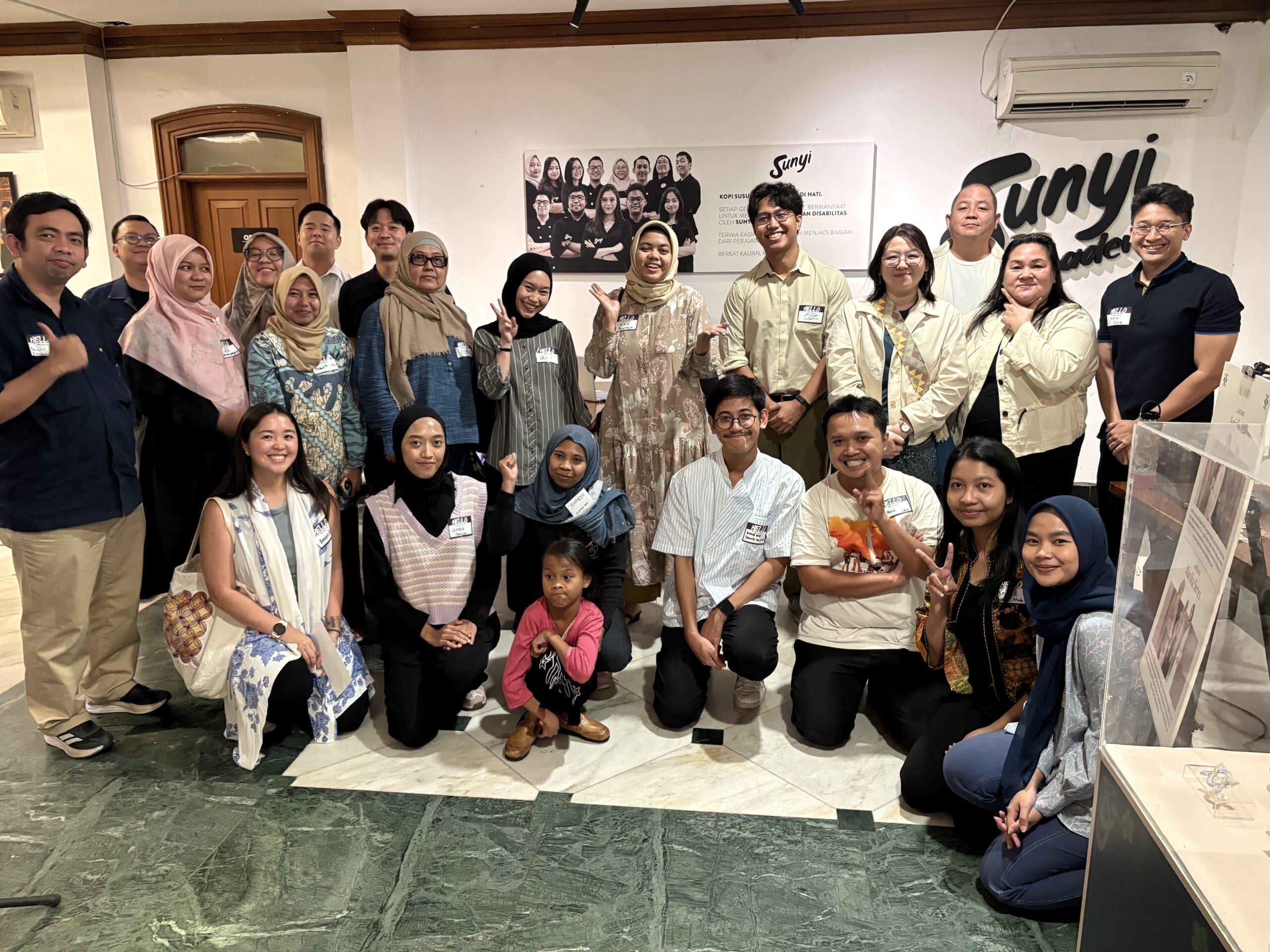 A group photo of participants standing and kneeling together inside the Sunyi Academy space. The diverse group smiles at the camera, with some making peace signs, wearing casual to semi-formal outfits and name tags. A Sunyi Academy logo and team photo are visible on the wall behind them, capturing a warm, inclusive, and community-driven atmosphere at Elevate's Focus Group Discussion as part of their needs assessment phase.